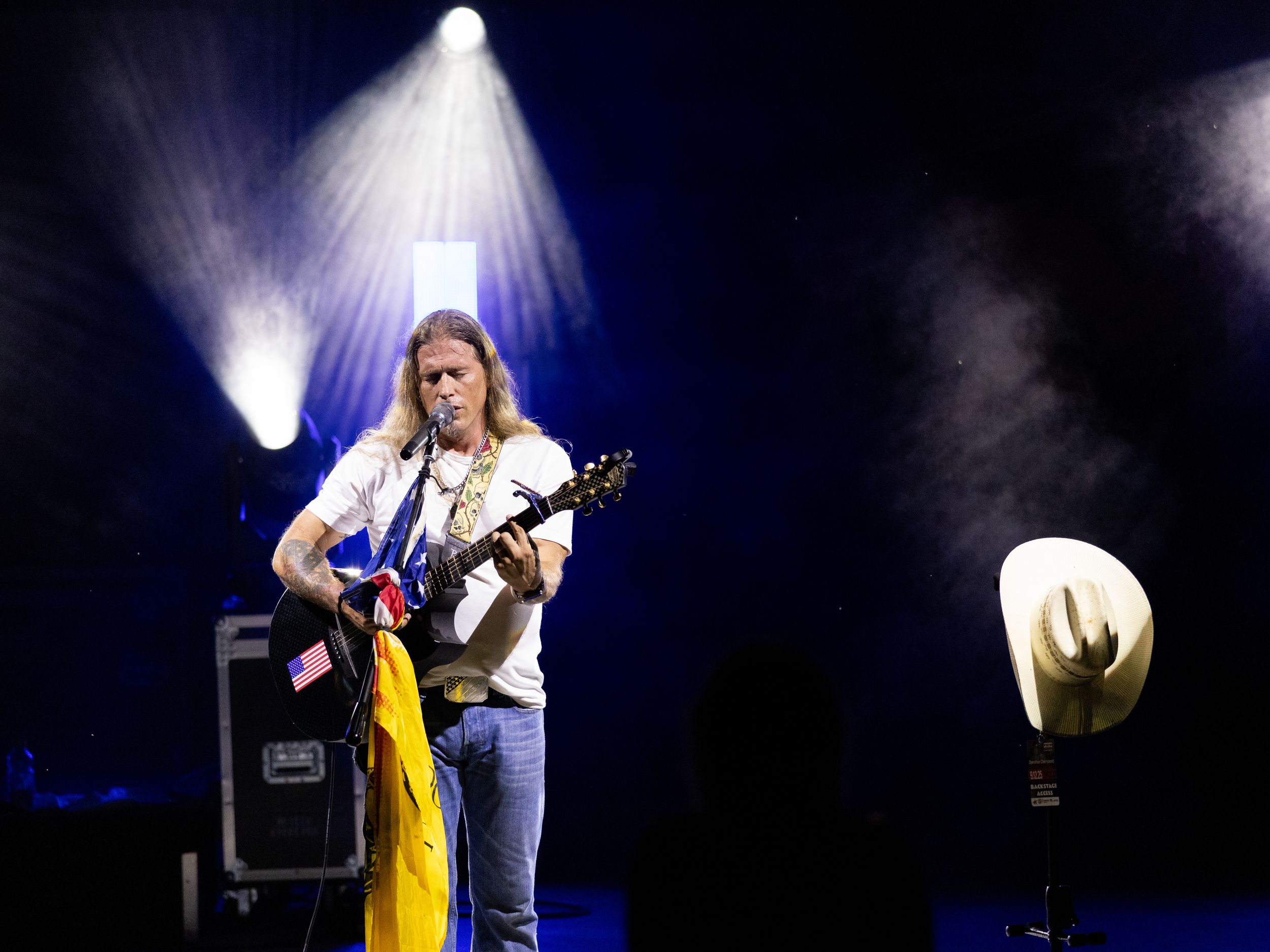 Musician playing electric guitar on stage under dramatic spotlights with a white cowboy hat and colorful flag nearby.