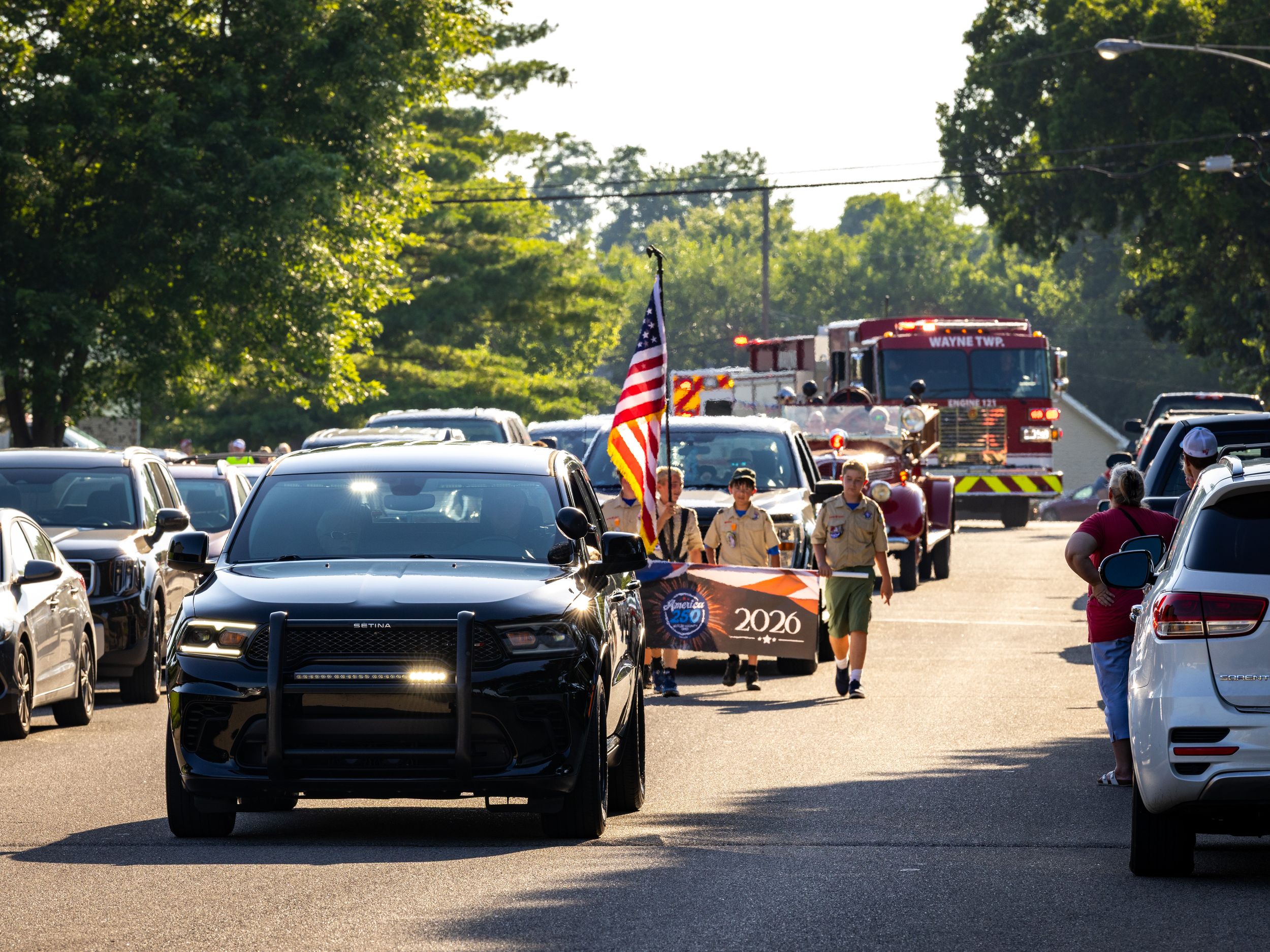 Police car leads a patriotic parade with fire truck and American flag on a tree-lined street.