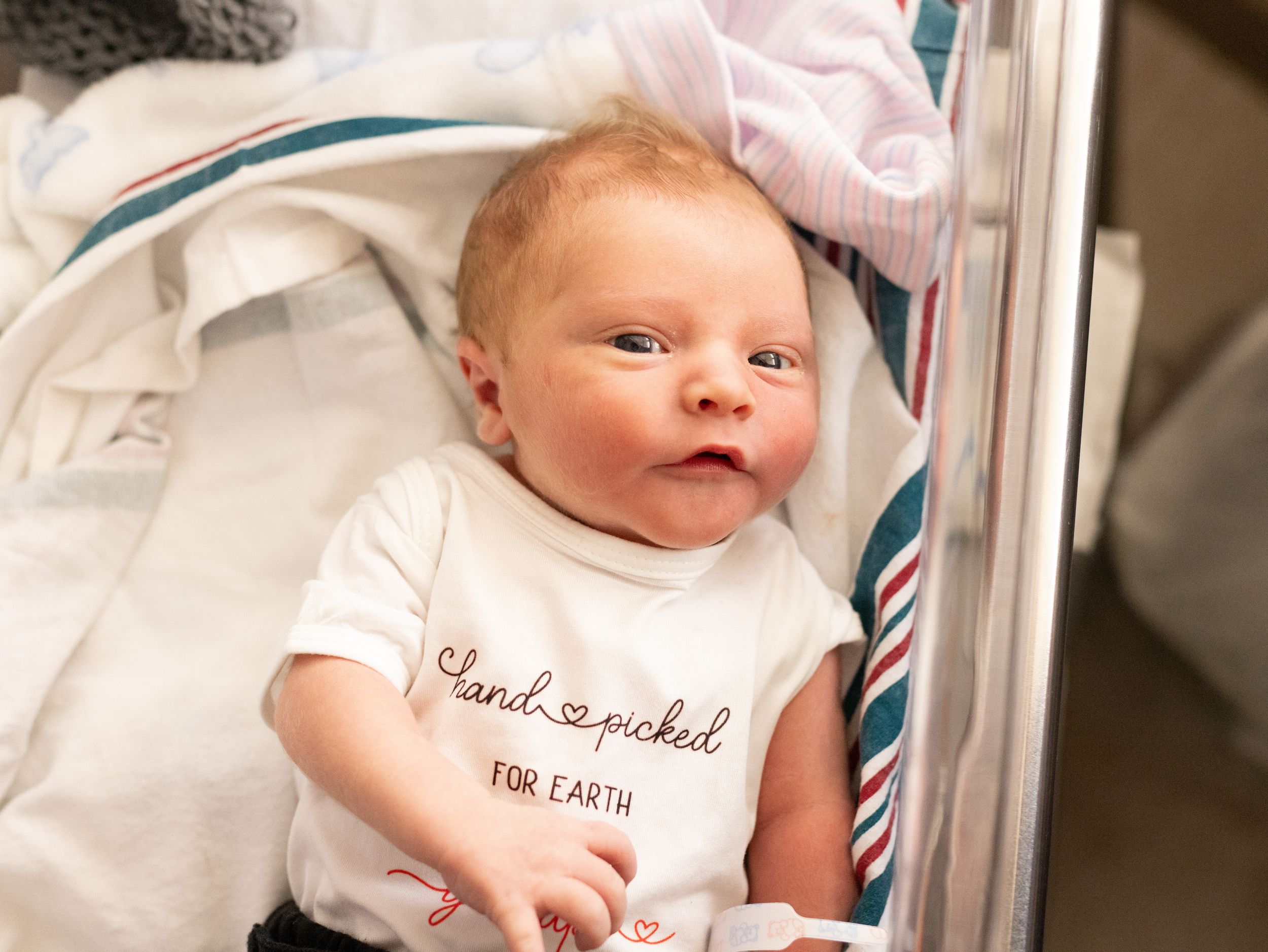 Newborn baby in white onesie reading hand-picked for Earth lying in hospital bassinet with striped blanket.