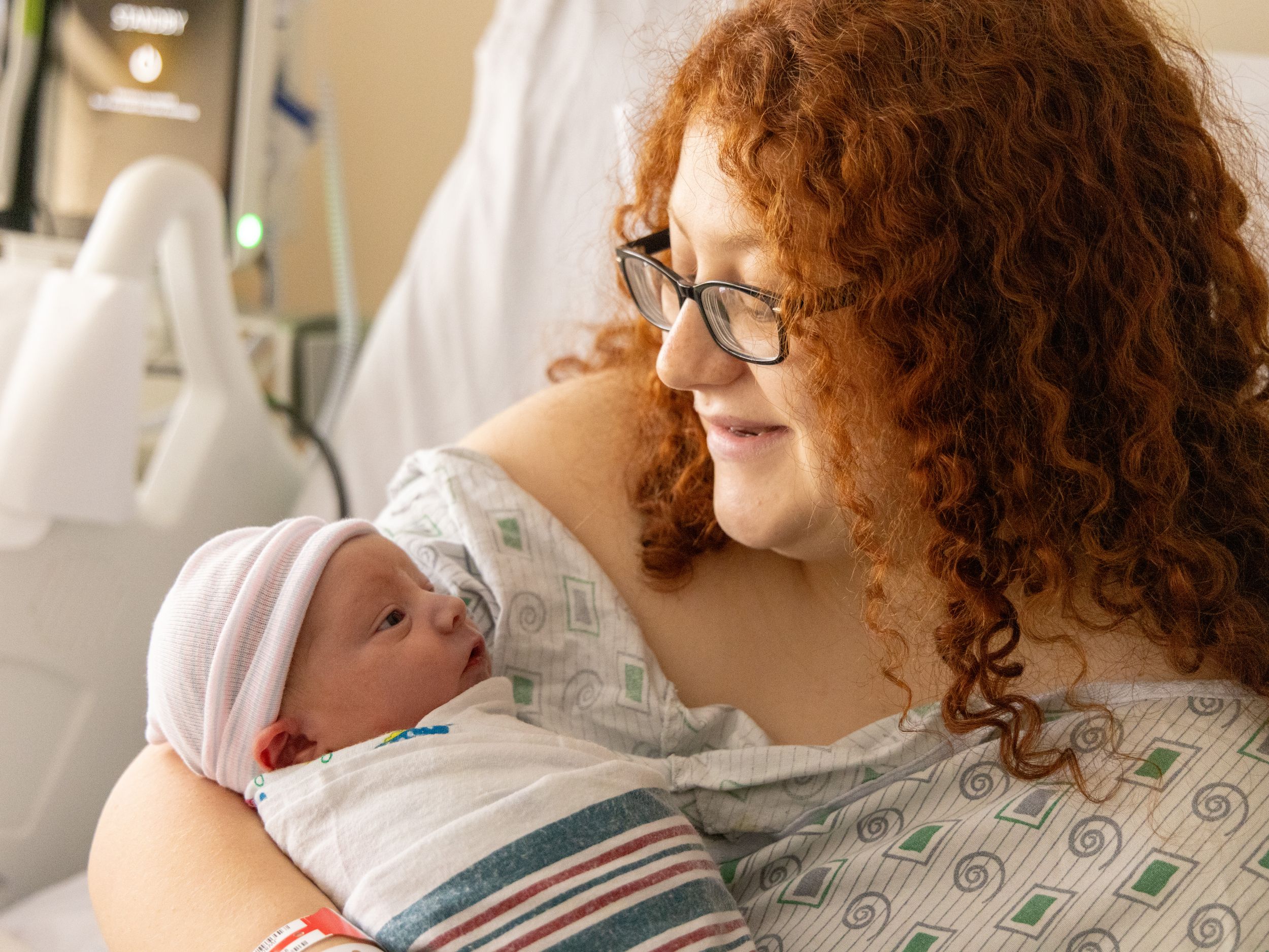 A red-haired mother wearing glasses gazes lovingly at her newborn baby in a hospital room.