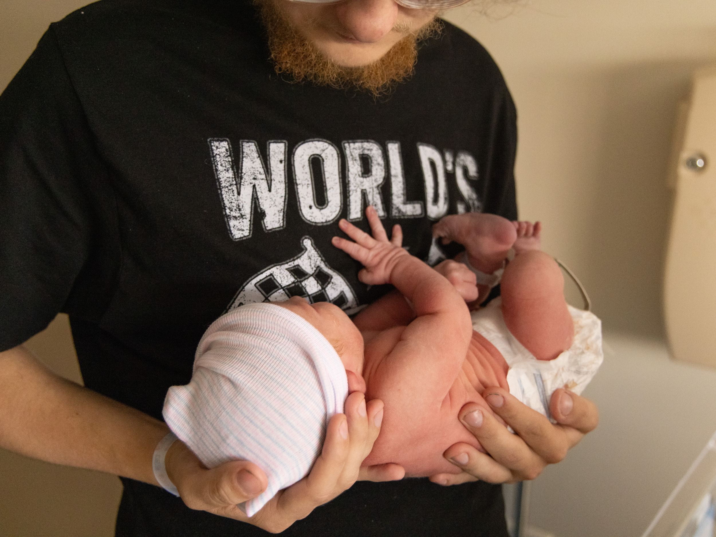 Father in World's Best Dad shirt proudly holds two newborn twins in his arms.