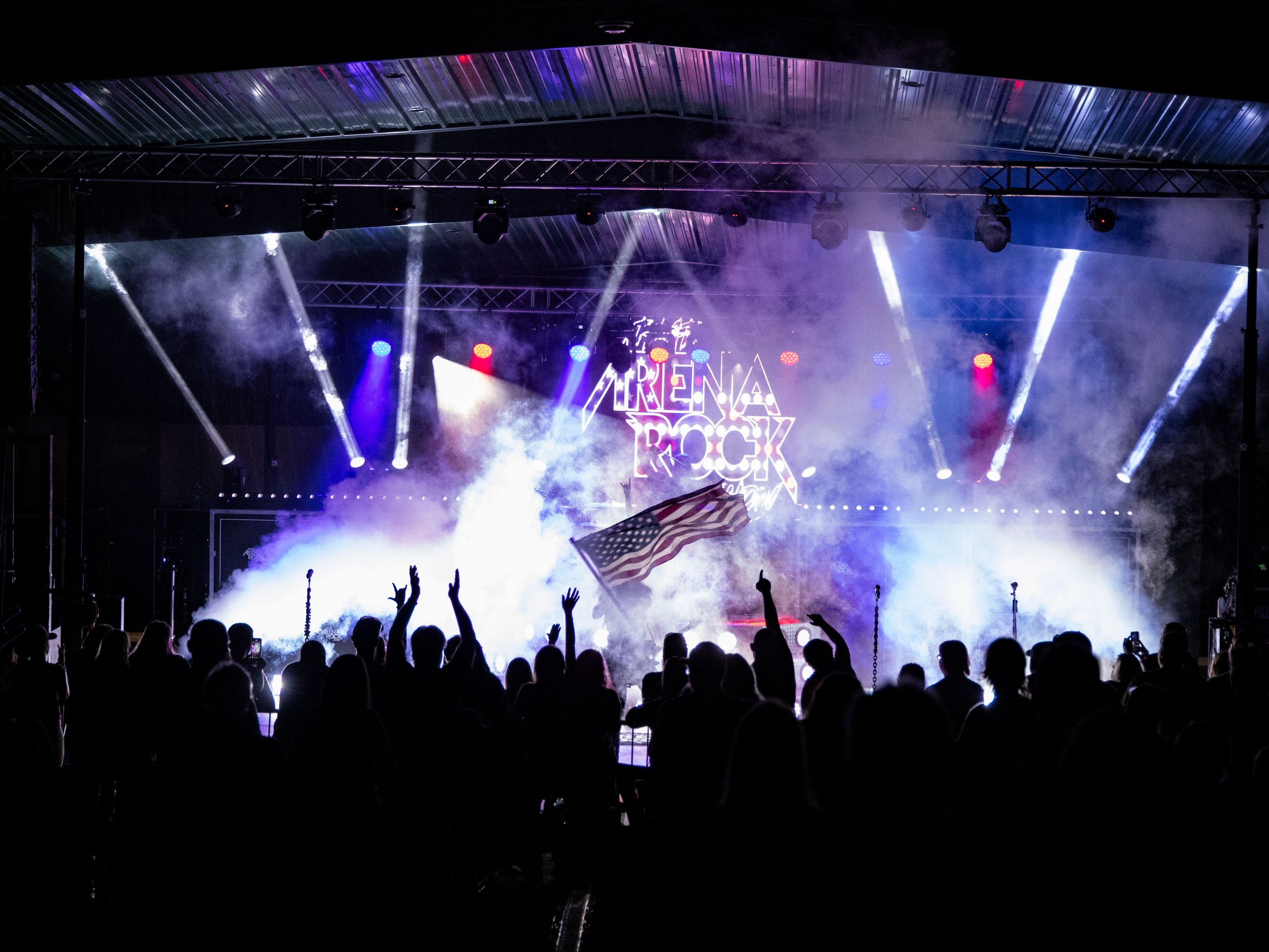 Crowd at an outdoor rock concert with dramatic stage lighting and smoke effects at night.