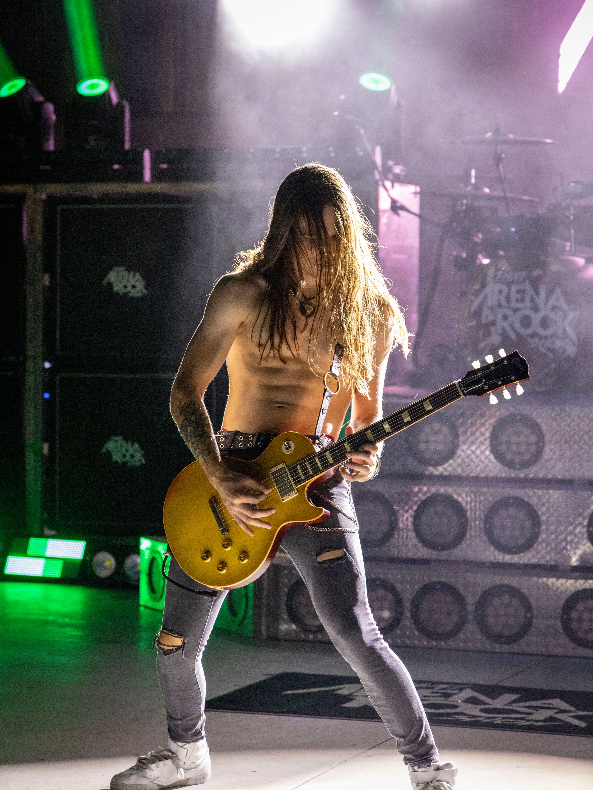 Shirtless male guitarist with long hair plays a gold Les Paul guitar on stage at an Arena Rock concert.