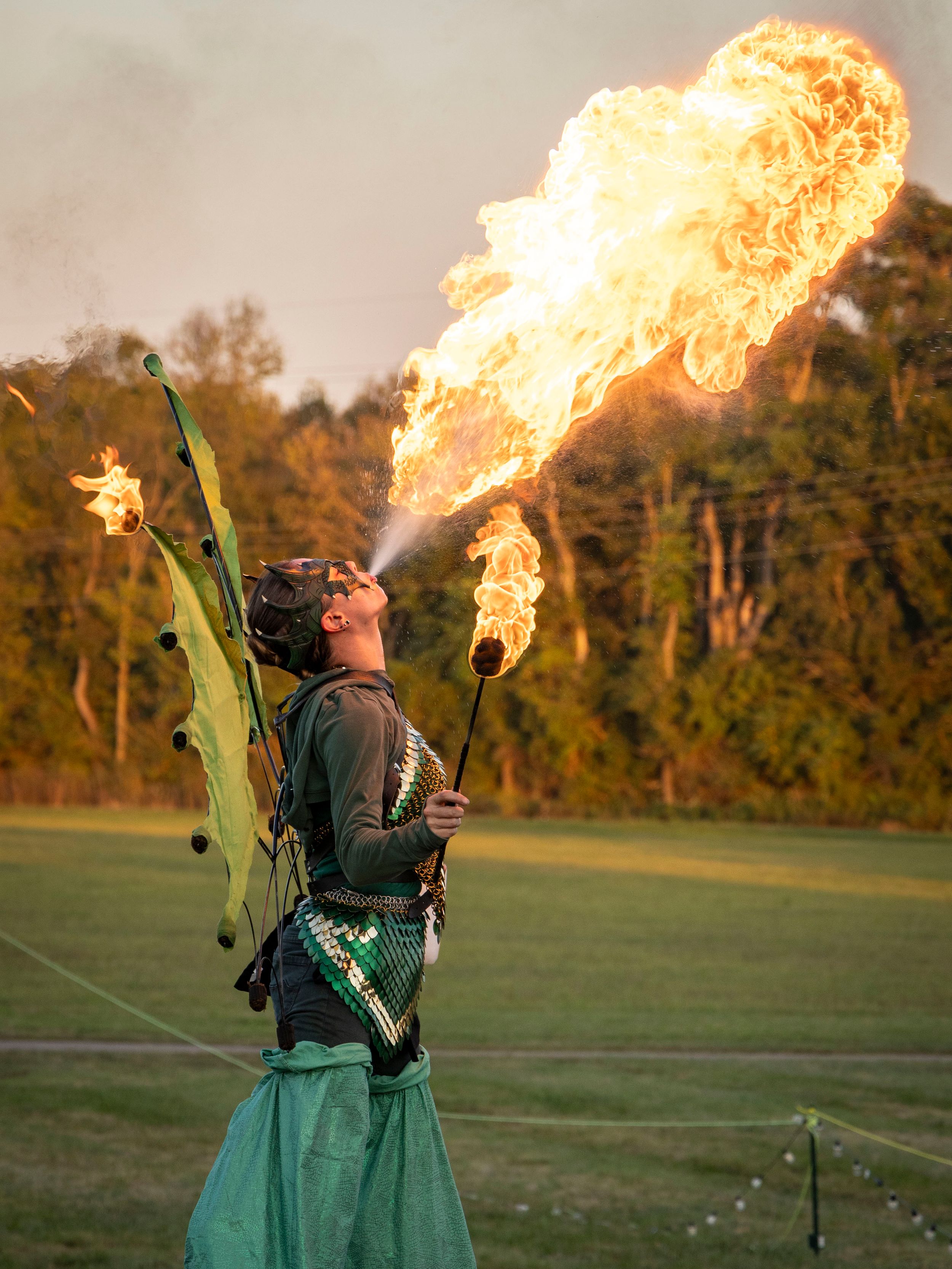 Fire breather in dragon costume performing at outdoor festival, exhaling large flames against a green field backdrop.
