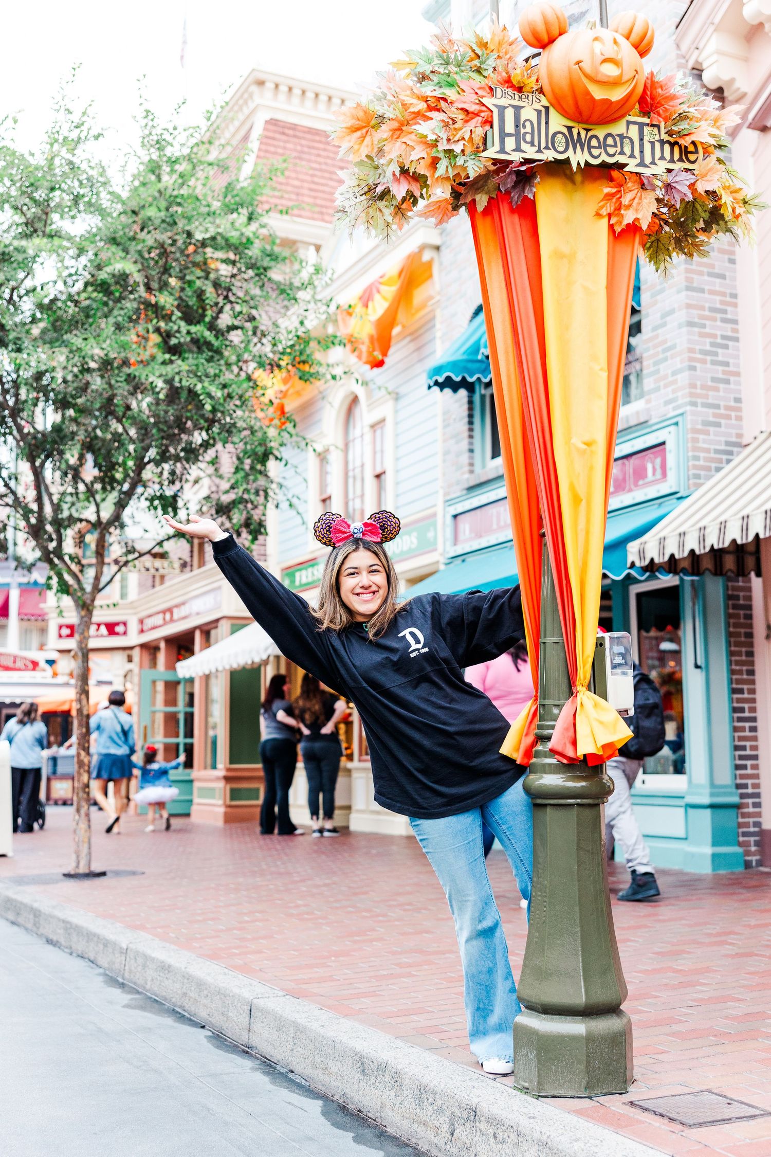 Enthusiastic pose at Disneyland wearing mouse ears and black sweatshirt near decorated lamppost.