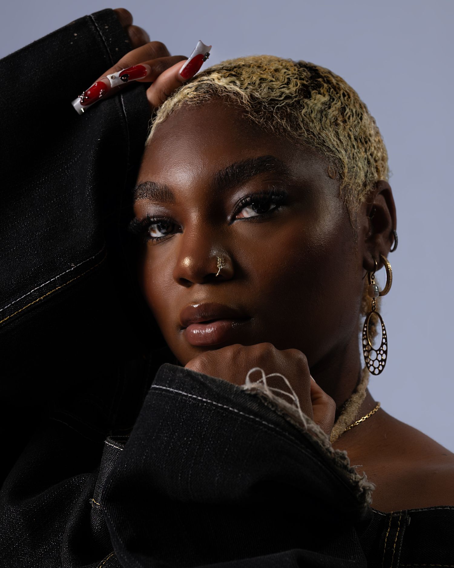 Portrait of elegant figure in black jacket with bleached blonde hair, nose ring, and dramatic lighting against gray background.