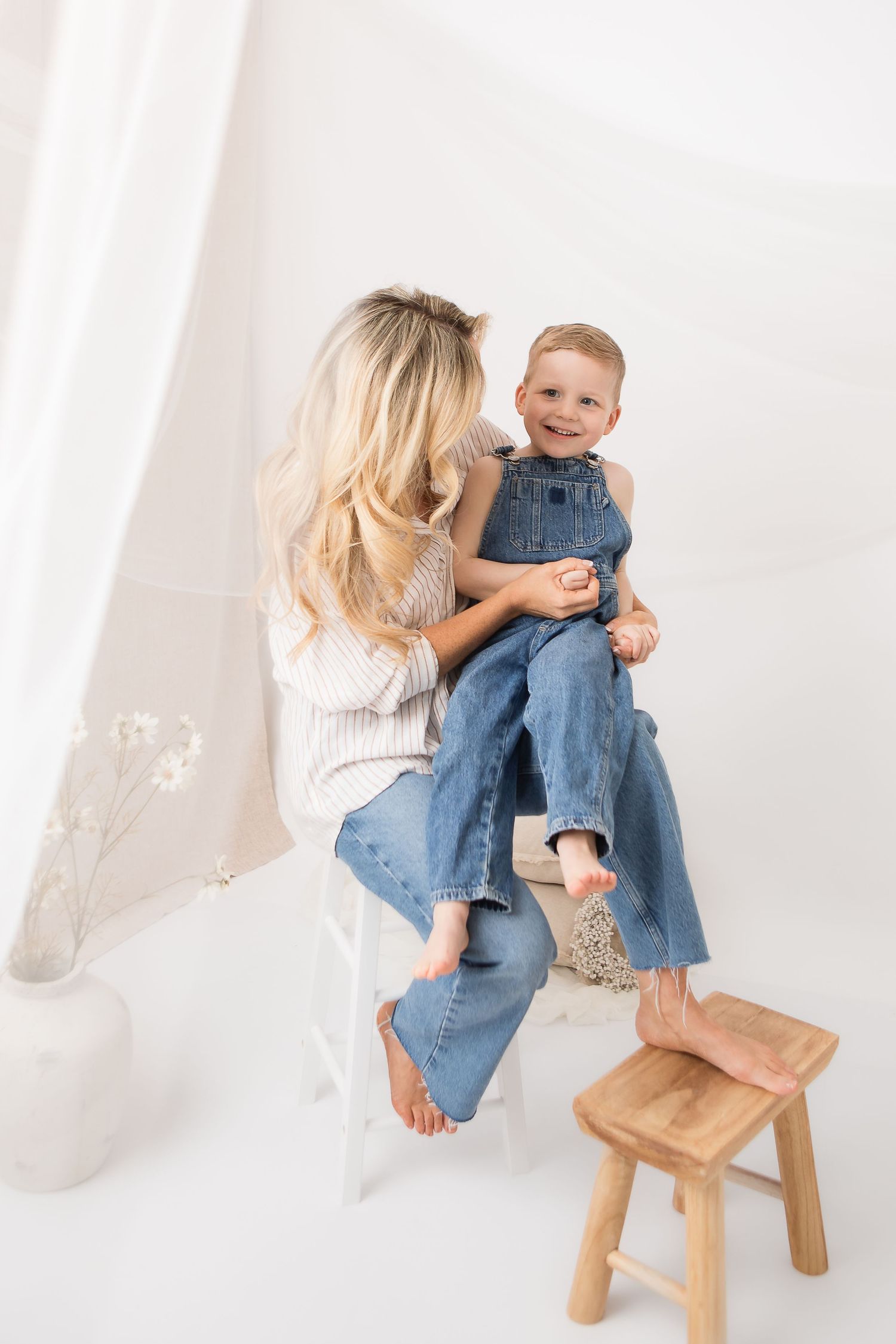 Two people sitting together in casual denim and white sweater in minimalist white studio setting with wooden stool.