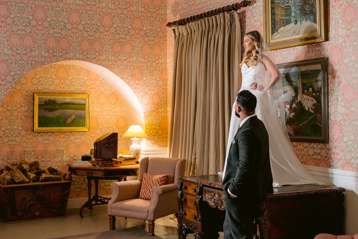 Bride standing on the table and groom holding her hand. Both smiling. Pearl veil. Black suit. Private castle. Vintage room. Ireland.