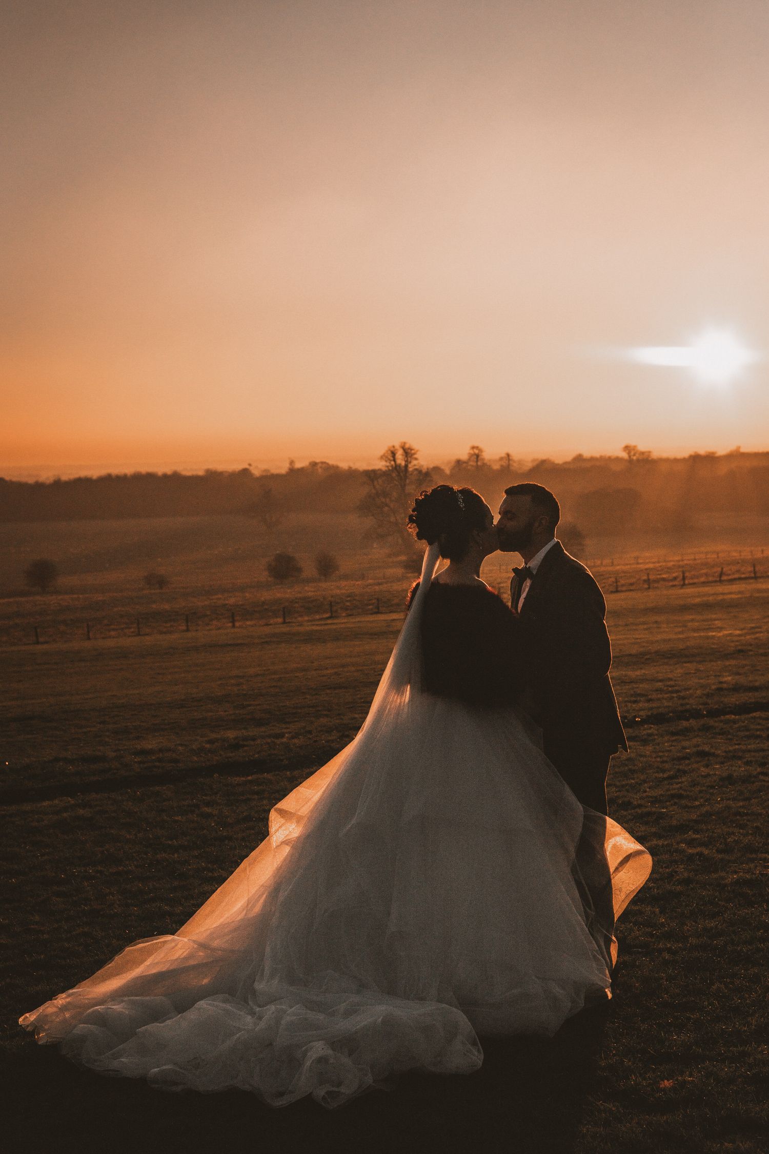 A silhouetted couple in wedding attire stands together against a warm sunset sky over rolling hills and fields.