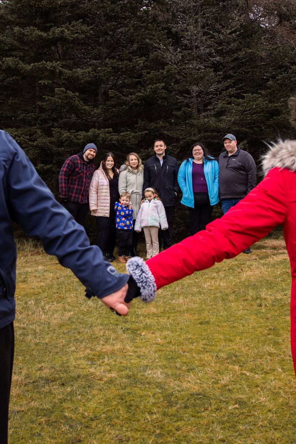 A group stands outdoors on grass while hands in the foreground form a heart shape against an evergreen forest backdrop.
