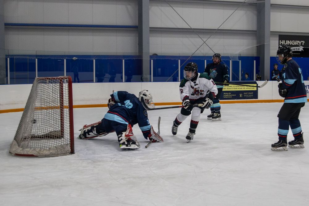 Ice hockey players in white and blue jerseys compete near the goal net during a game at an indoor rink.