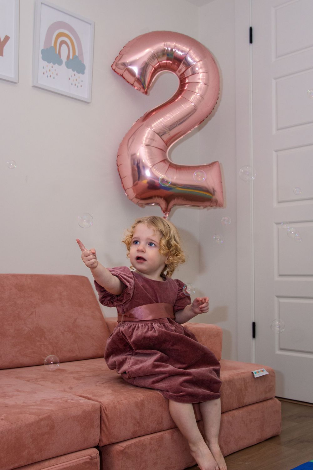 A large rose gold number 2 balloon floats above a dusty pink couch where a small child in a mauve dress sits excitedly pointing at bubbles.