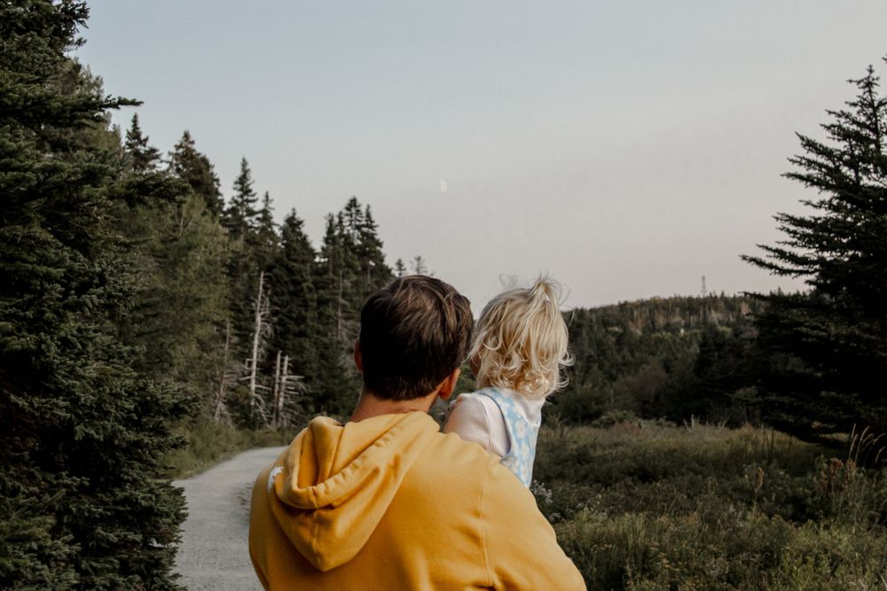 Dad & Daughter on a trail in the woods, trees on the left, crescent moon in the middle. Daughter is pointing at the moon.