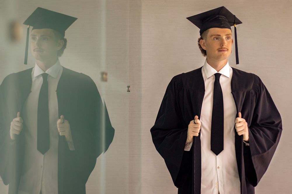 A graduate in black cap and gown with a black tie stands next to their shadow reflection on a white wall.