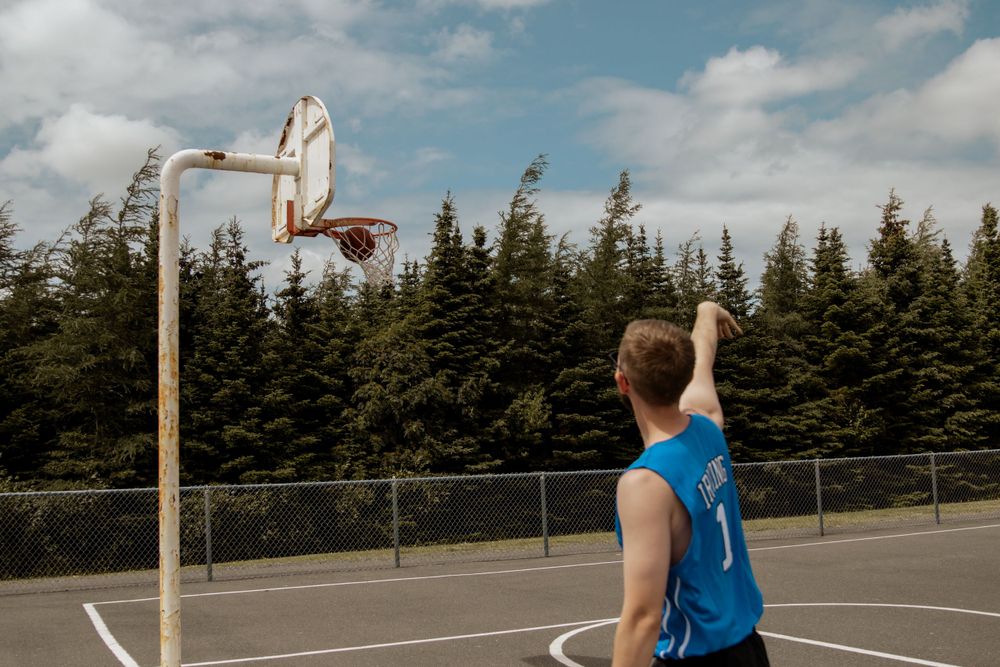 Basketball player in blue jersey practices shooting on an outdoor court surrounded by evergreen trees under blue sky.