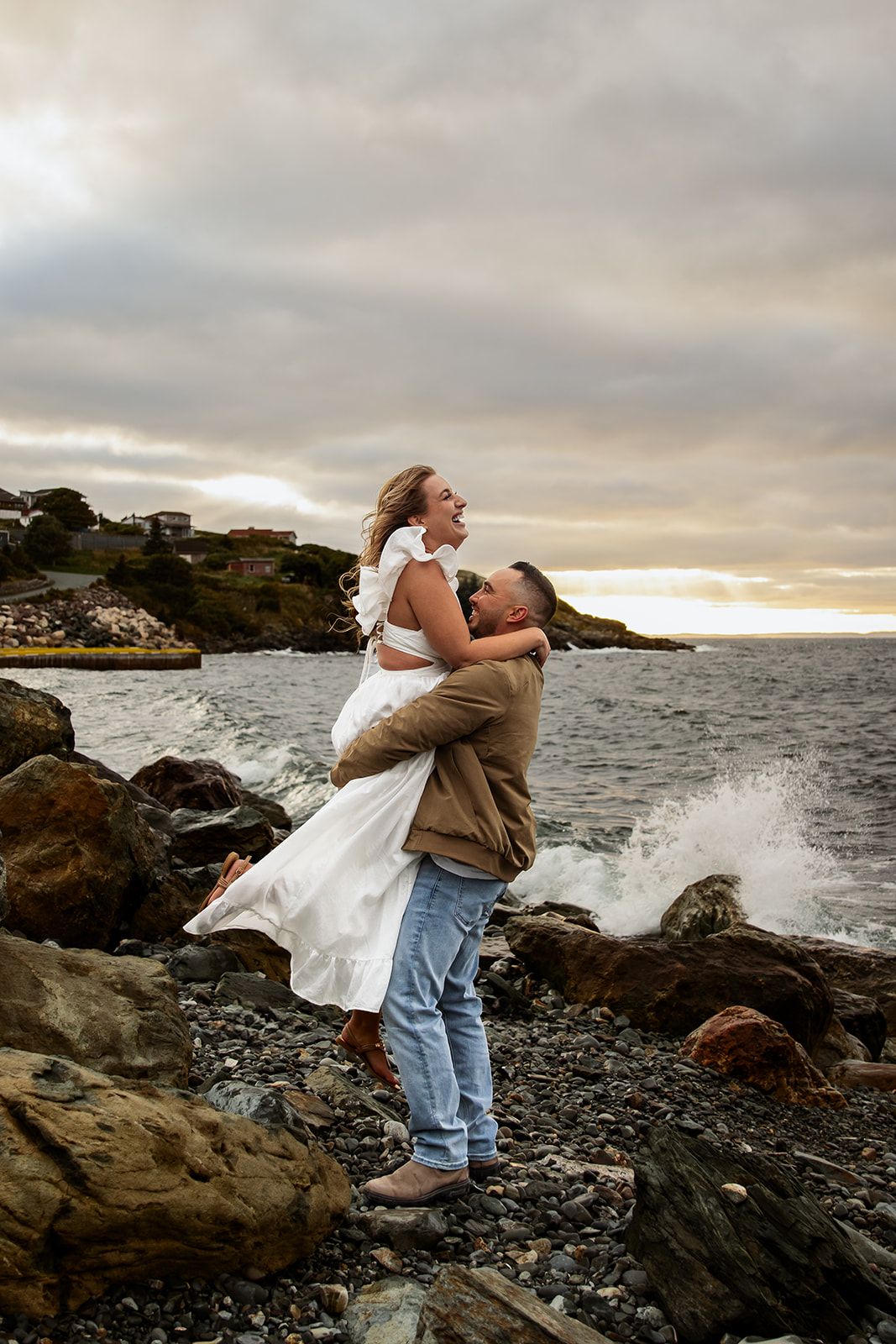 A couple embraces on rocky coastal cliffs at sunset as waves crash against the shore.