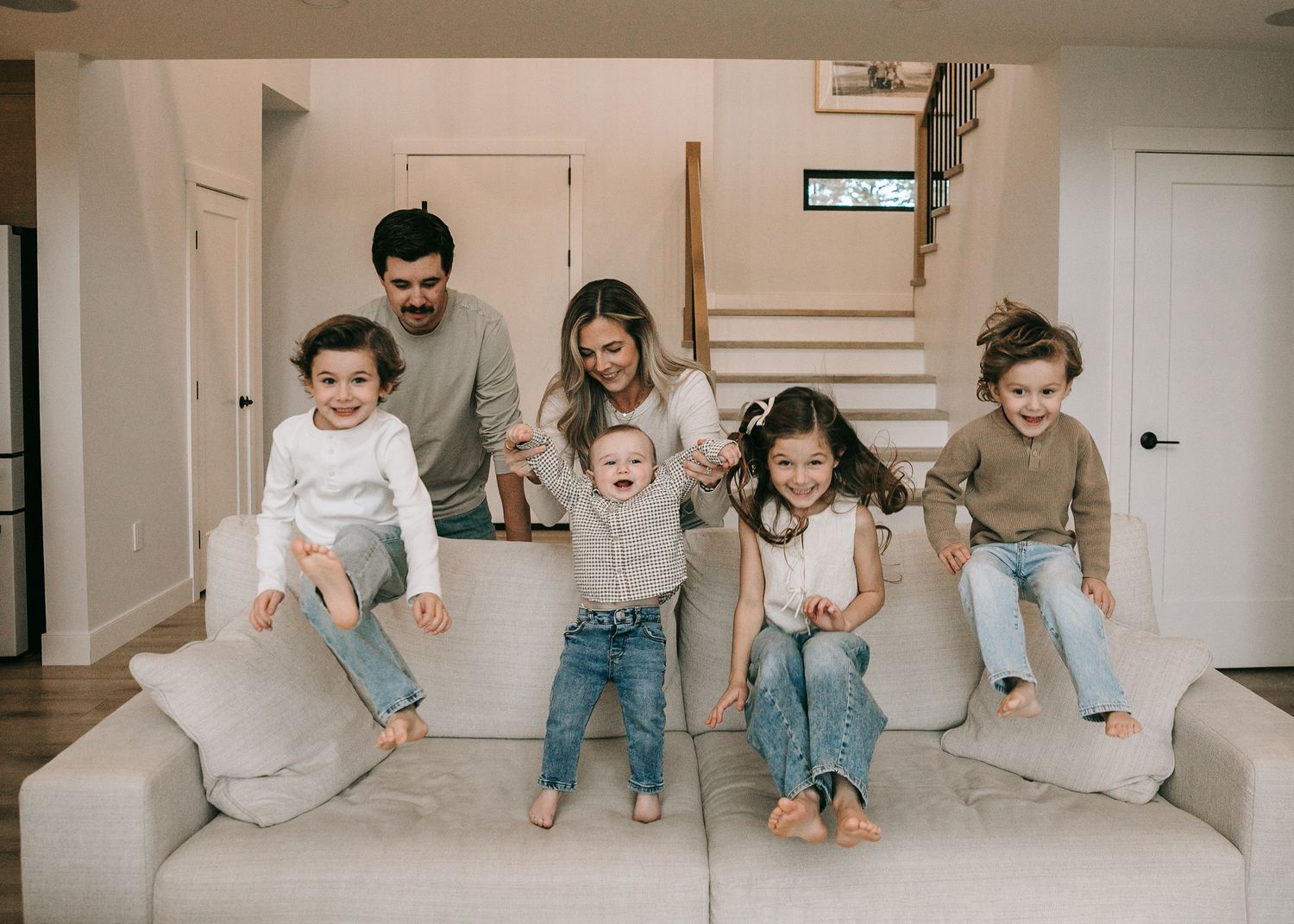 Energetic family with four young children jumping on a white couch in a modern bright home.