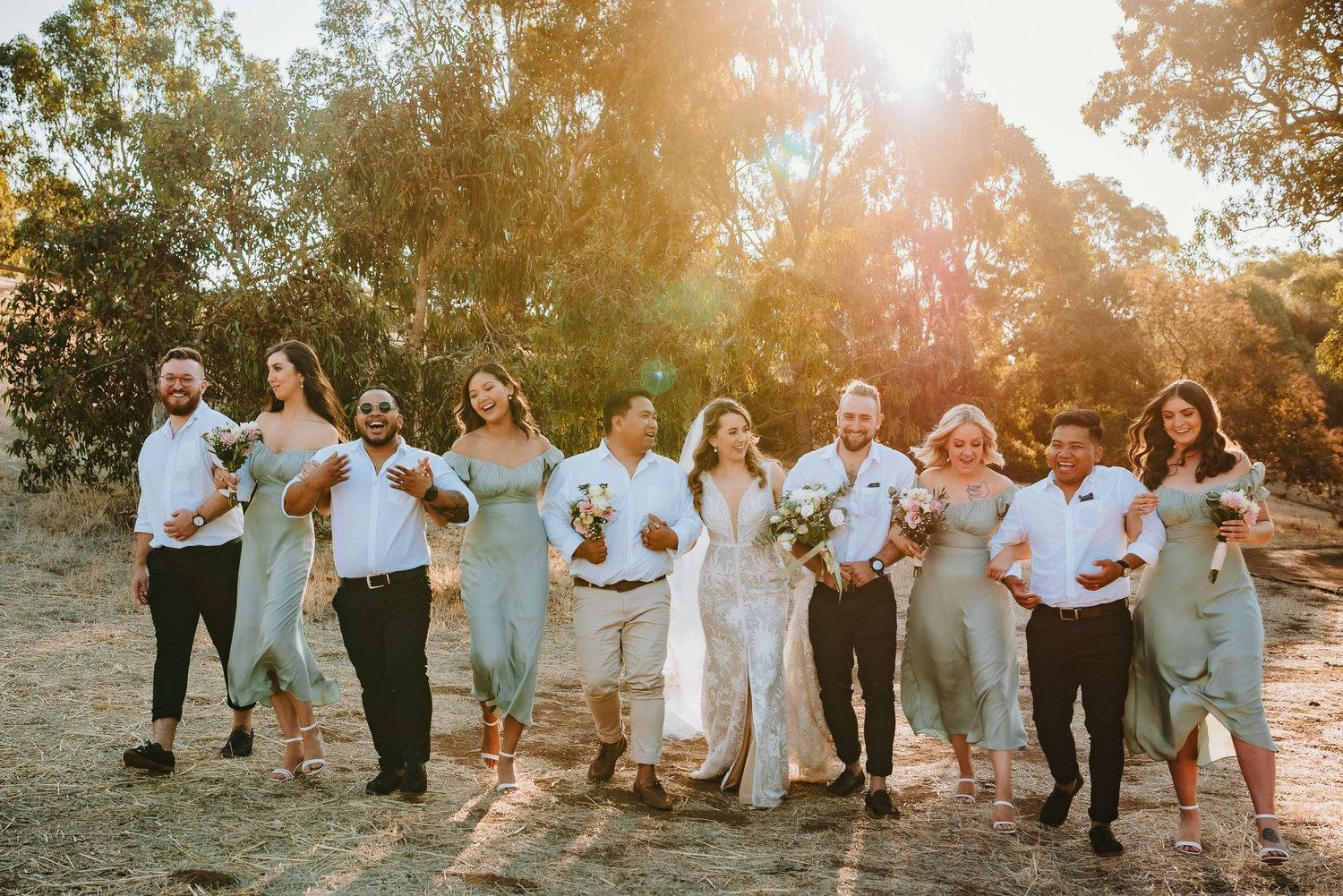 A group of wedding party members walk arm in arm during sunset in a scenic outdoor setting.