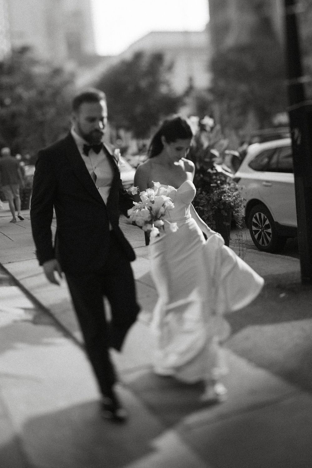 A black and white photo of a bride and groom walking together on a city sidewalk, with the bride holding a bouquet.