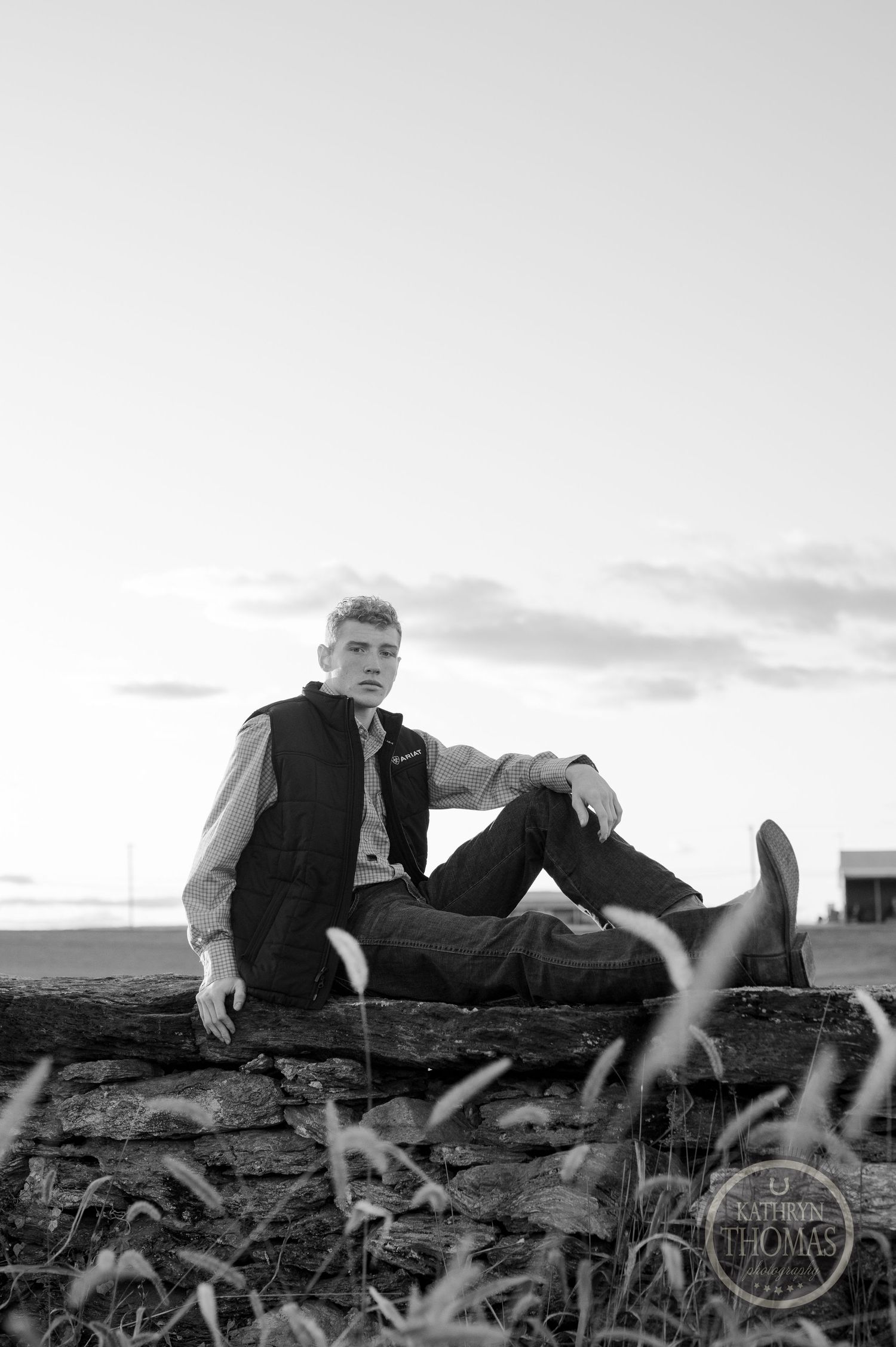 Black and white portrait of person sitting on stone wall with grass in foreground.