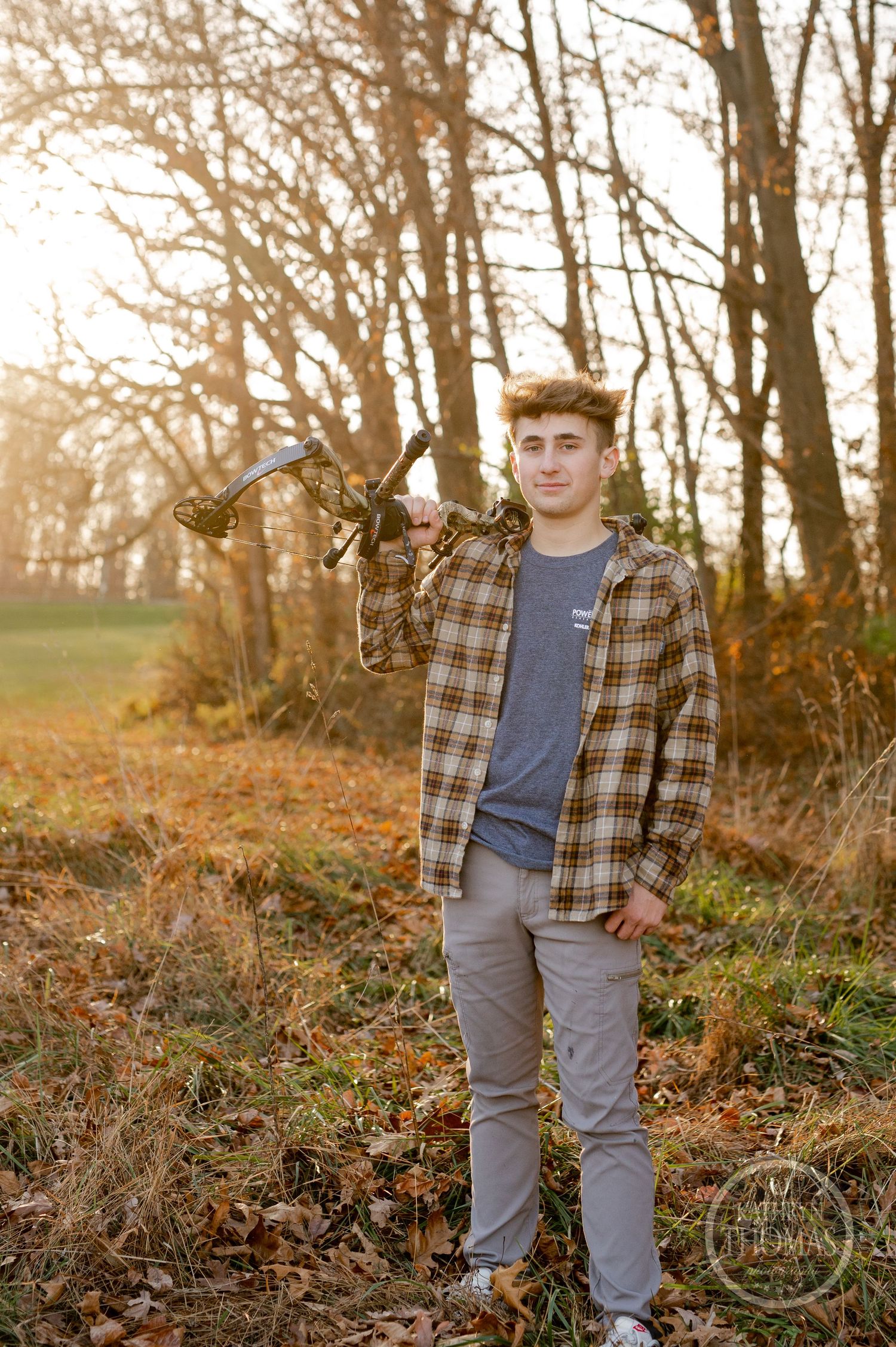 Archer in plaid flannel shirt poses with compound bow in autumn forest at golden hour.