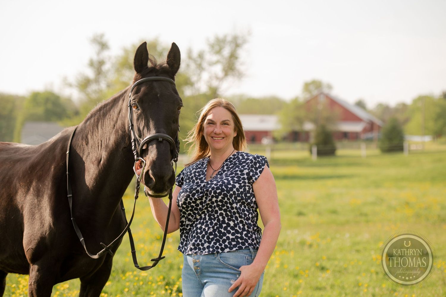 A black horse and person stand in a green field with farm buildings in the background.
