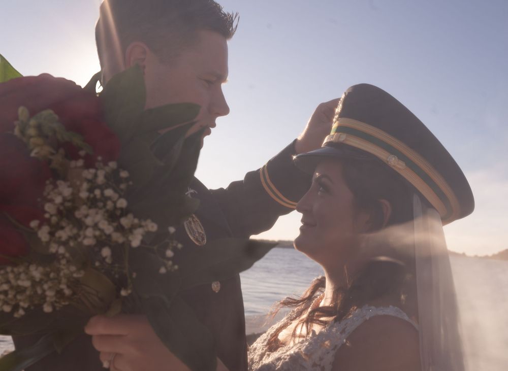 A couple embraces in soft sunlight during their lakeside military wedding celebration with flowers and a veil.