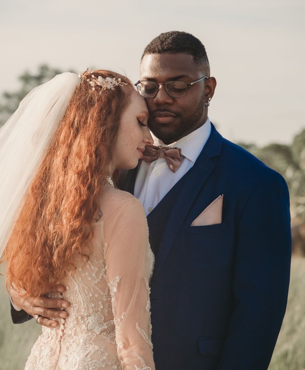 A romantic wedding portrait of a couple embracing outdoors, with the bride in lace and groom in navy suit with bowtie.
