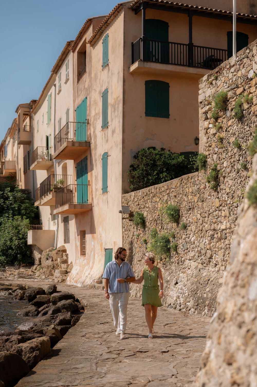 Couple walking along a charming cobblestone street with rustic stone walls and turquoise shutters.