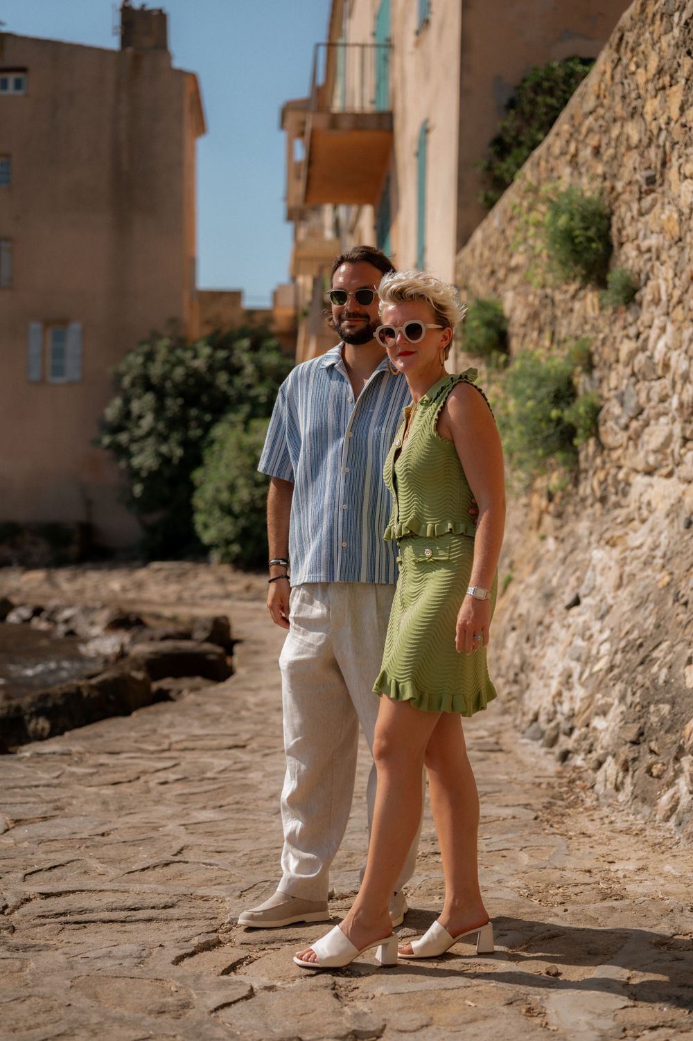 People strolling down a scenic stone pathway alongside Mediterranean-style buildings with teal shutters.