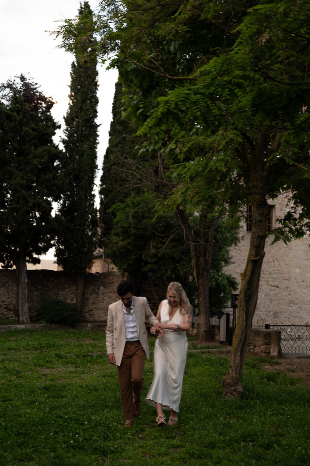 Couple walking through lush garden path with stone walls and cypress trees.