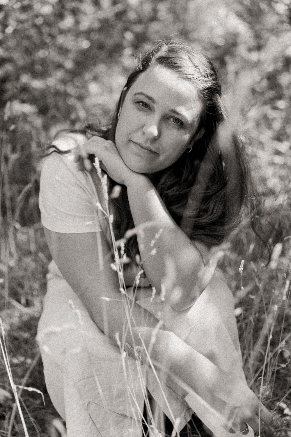 woman squatting in tall grass and looking at the camera with a soft face, black and white