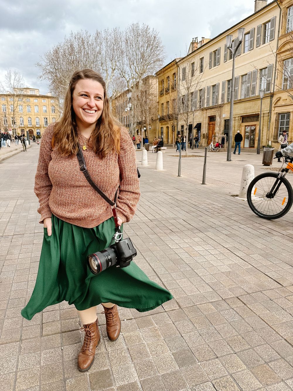 woman in Provence town square wearing a brown sweater, green skirt, and a camera