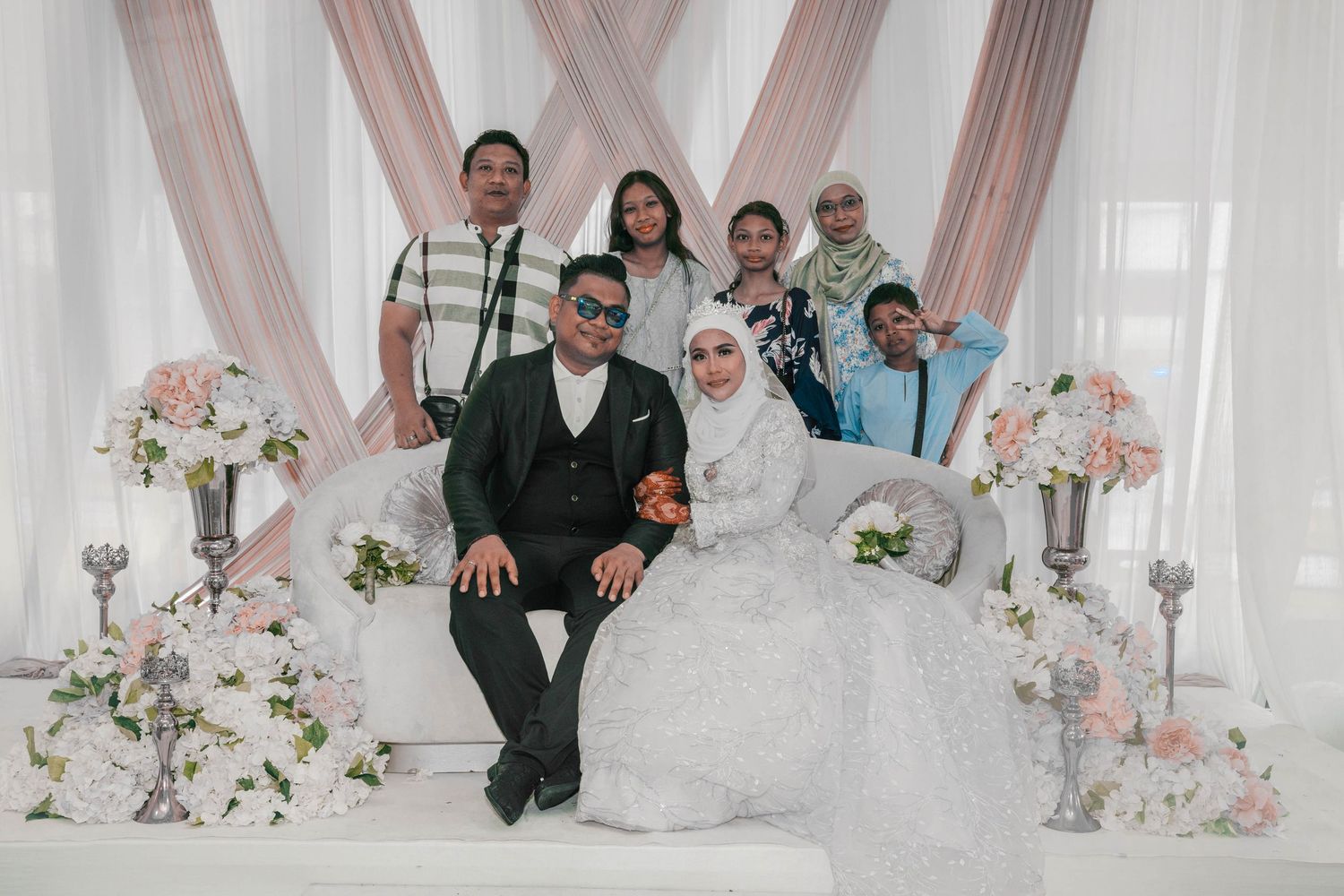 Wedding party group photo with family members against floral decorated white backdrop.