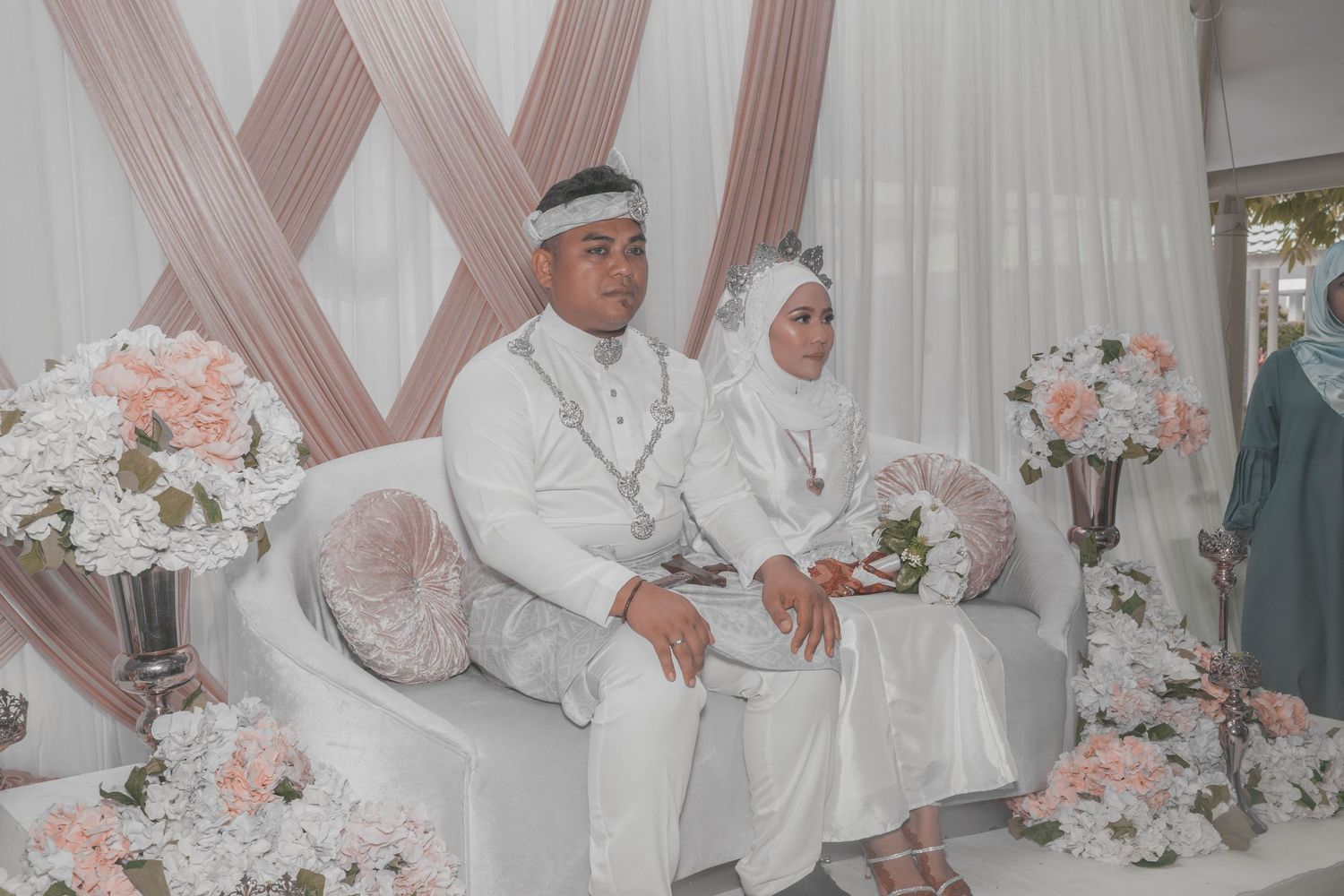 Muslim bride and groom in white attire sit together on ornate sofa decorated with pink and white floral arrangements.