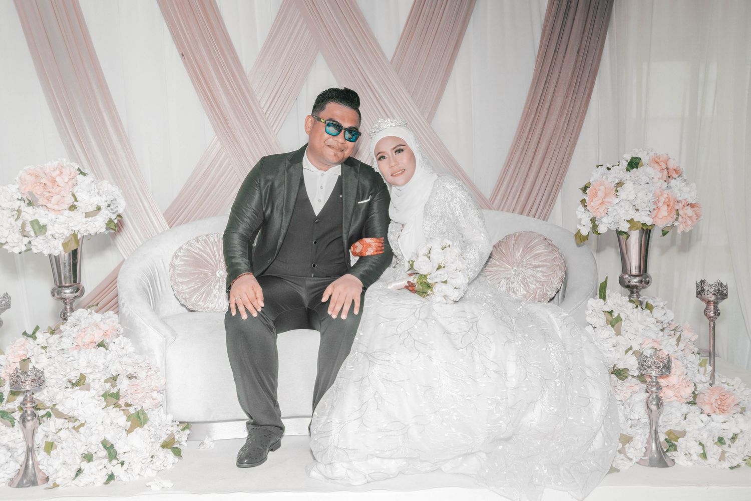 A wedding couple poses on an elegant white couch with floral decorations and draped fabric backdrop.