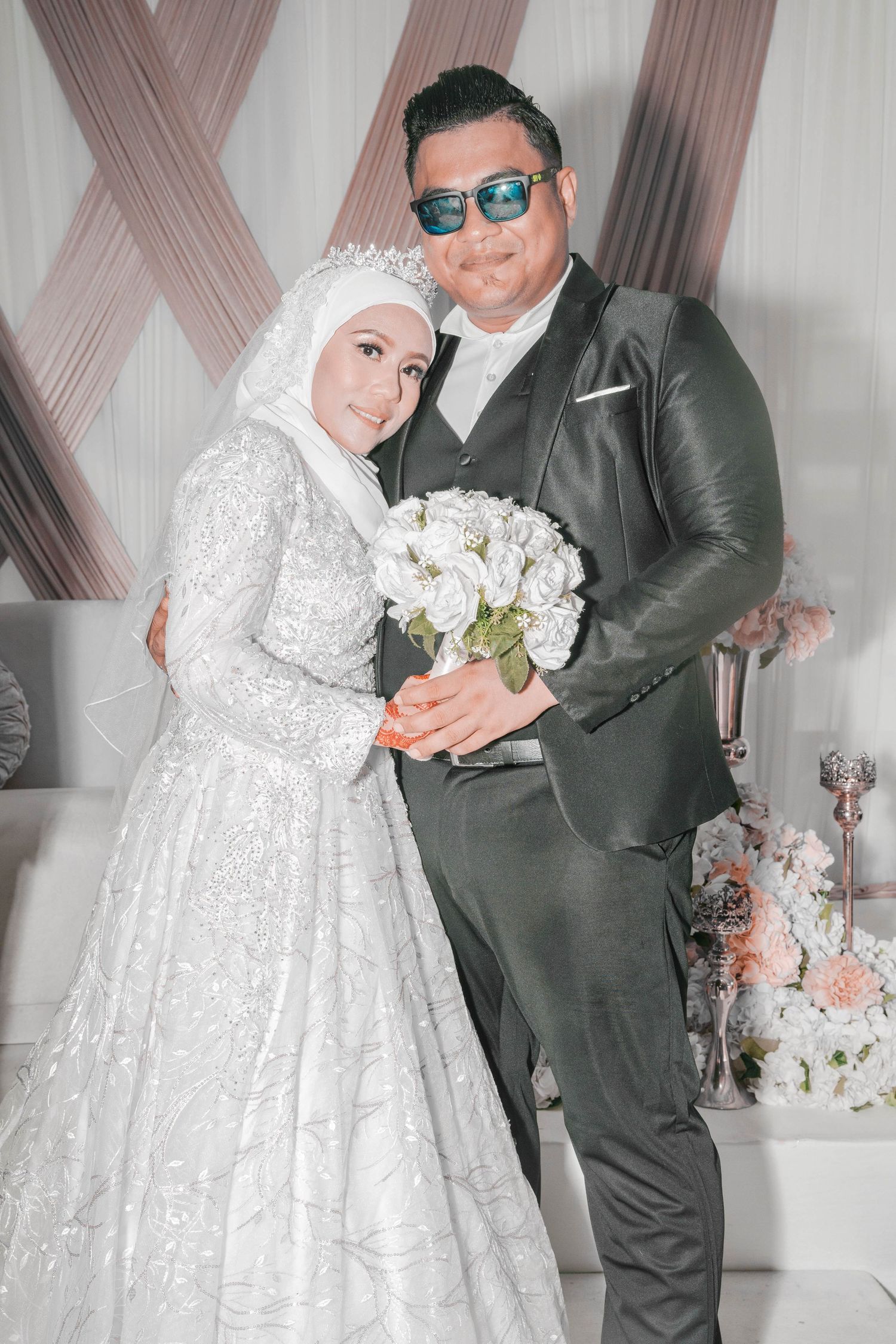 A bride in white lace gown and groom in gray suit hold a white bouquet against draped backdrop.