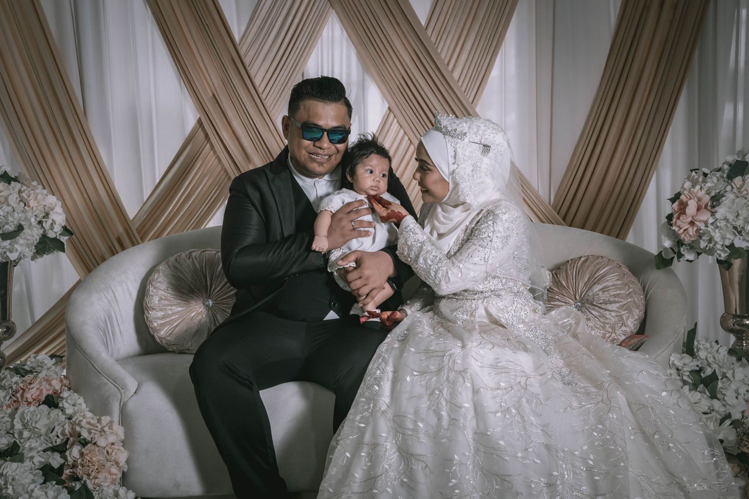 Wedding family portrait with couple and baby on elegant white sofa with floral decor.