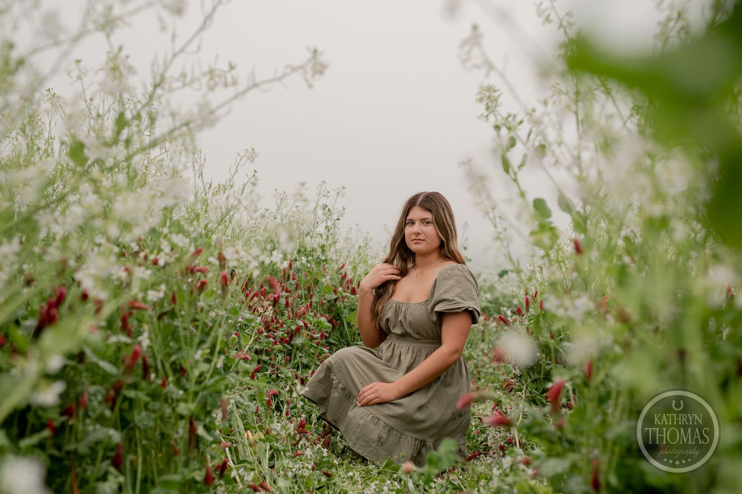 Someone in a green dress poses casually amid blooming wildflowers on a foggy day.