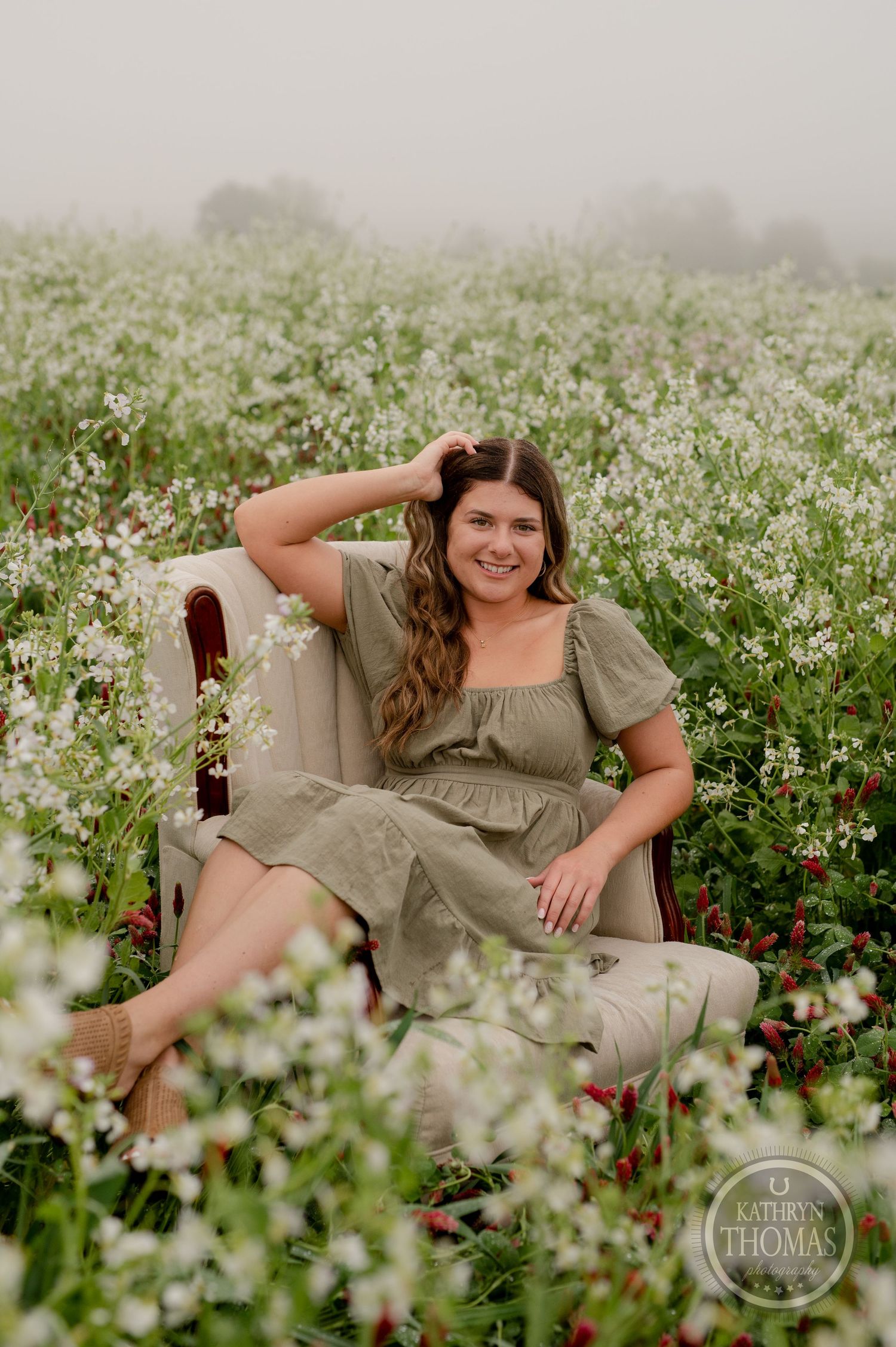 A woman poses casually in a plush armchair surrounded by white wildflowers in a misty field.