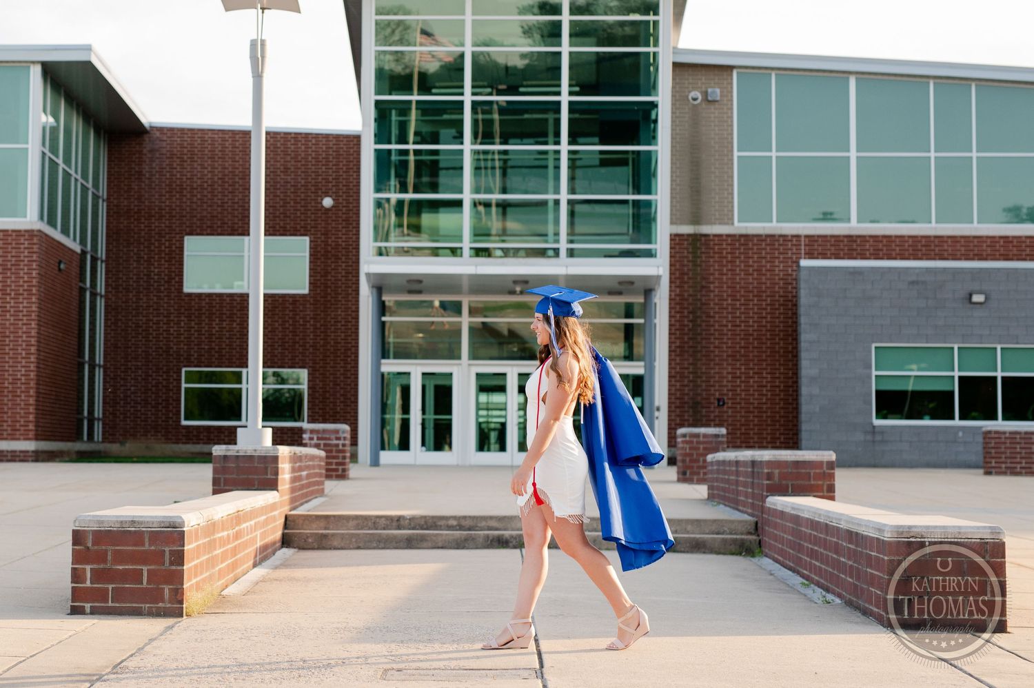 A graduate in a white dress and blue cap walks with a diploma outside a modern brick building.