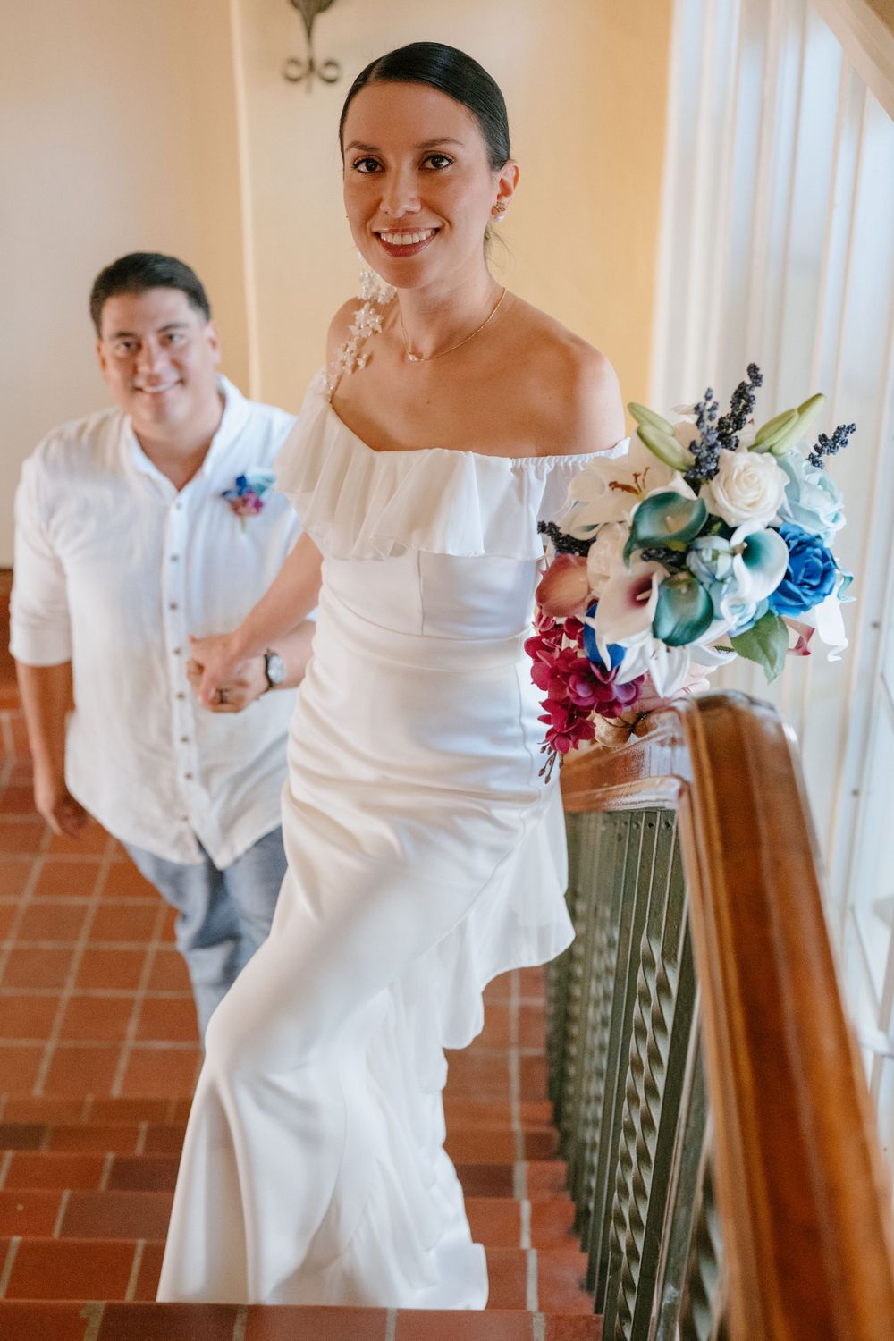 Elegant bride in off-shoulder white dress holds colorful bouquet on staircase.