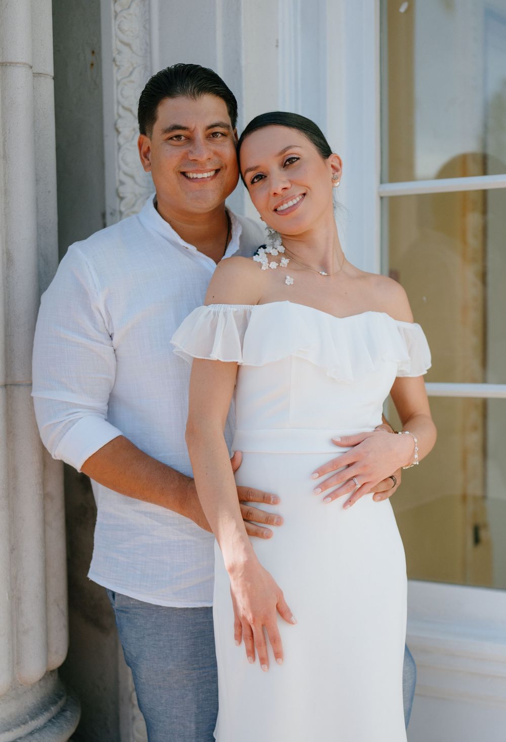 A newlywed couple poses against a white column in casual wedding attire.