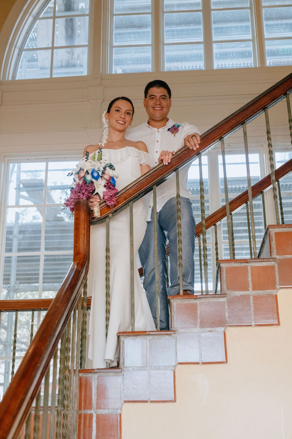 Wedding couple poses on elegant staircase with large arched window and natural light.