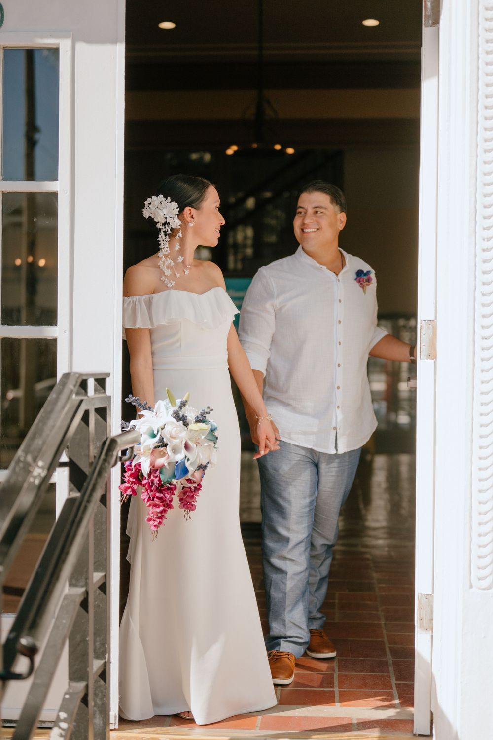 A bride holding a colorful bouquet stands with her partner in a doorway.