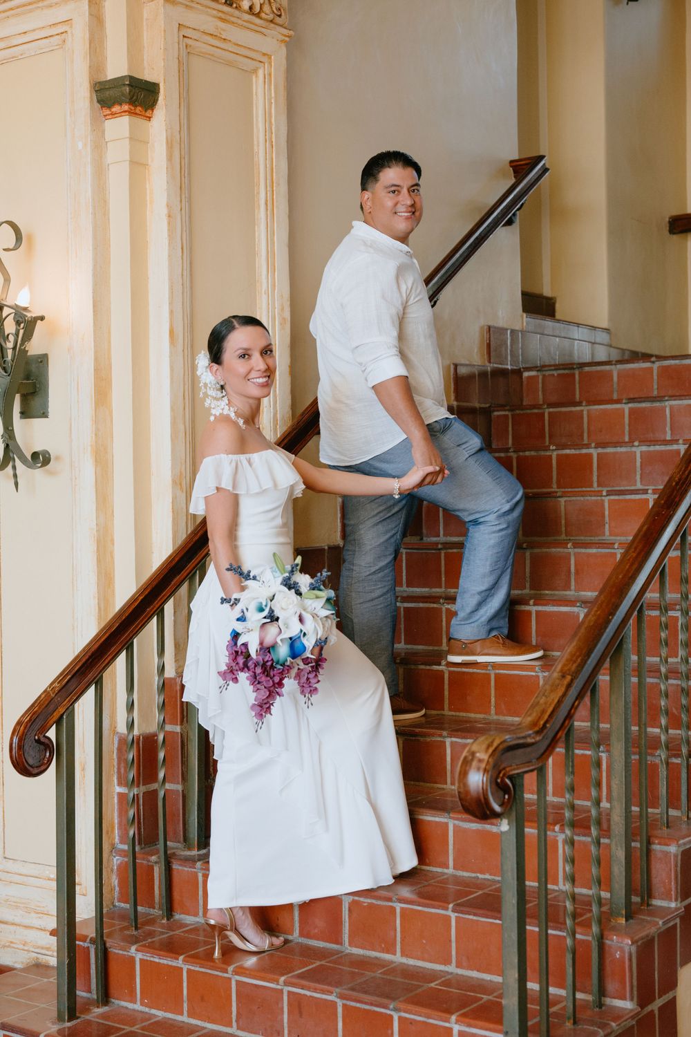 Wedding couple poses playfully on brick stairs with elegant white dress and bouquet.