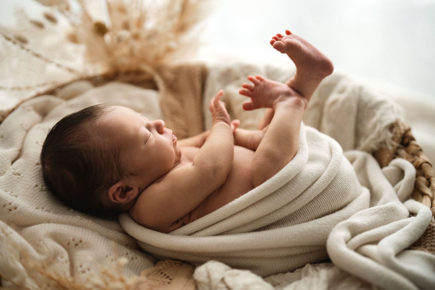 Newborn baby peacefully sleeping on cream colored linens wearing a white romper.