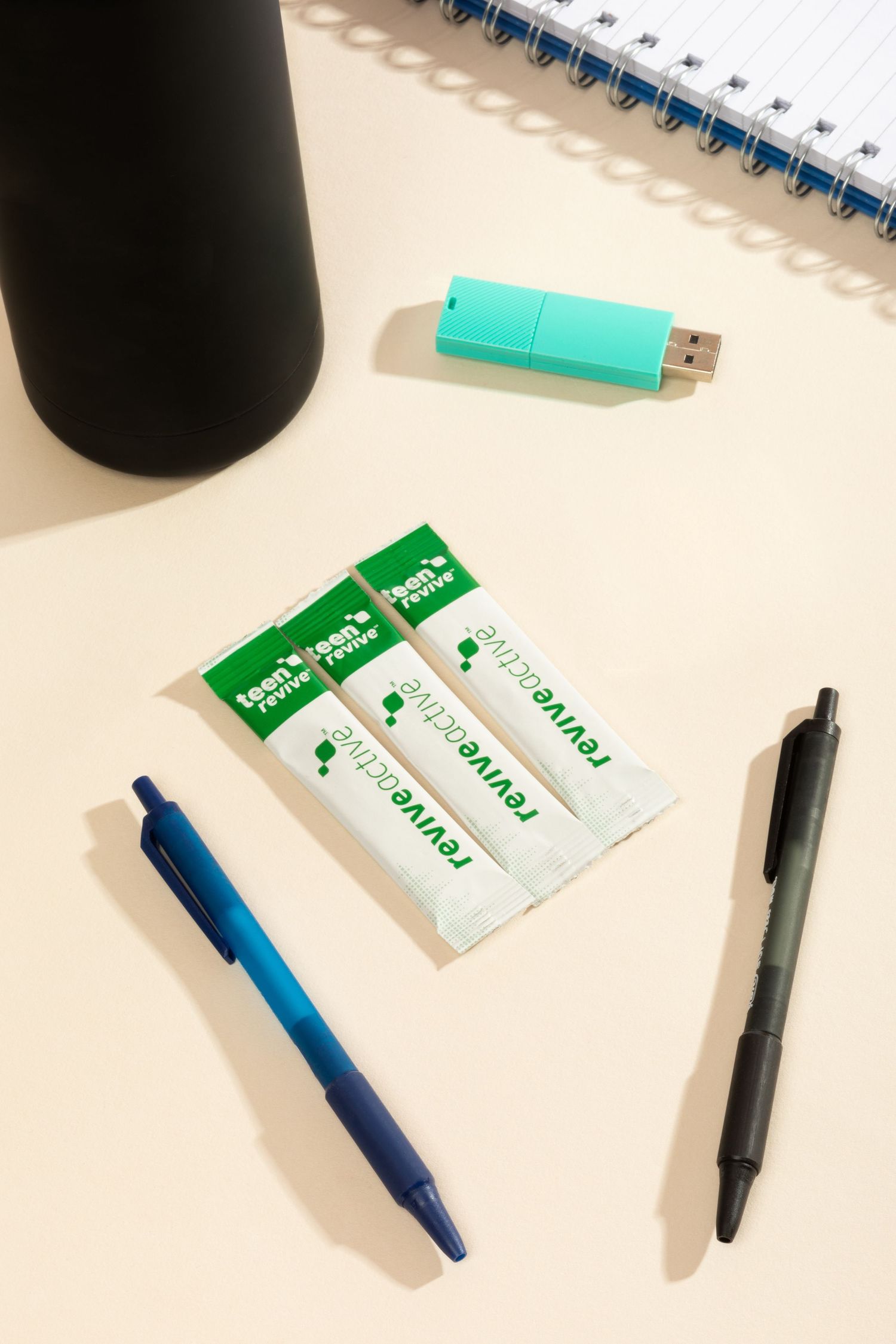 Office supplies arrangement with turquoise USB drive, pens, and white packets on desk surface. Three supplement sachets are also on the desk