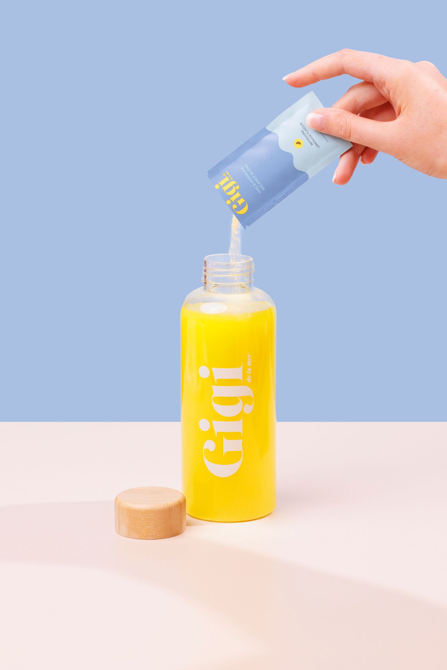 Pouring Gigi supplement powder from sachet into a glass branded bottle against pastel blue and beige background.