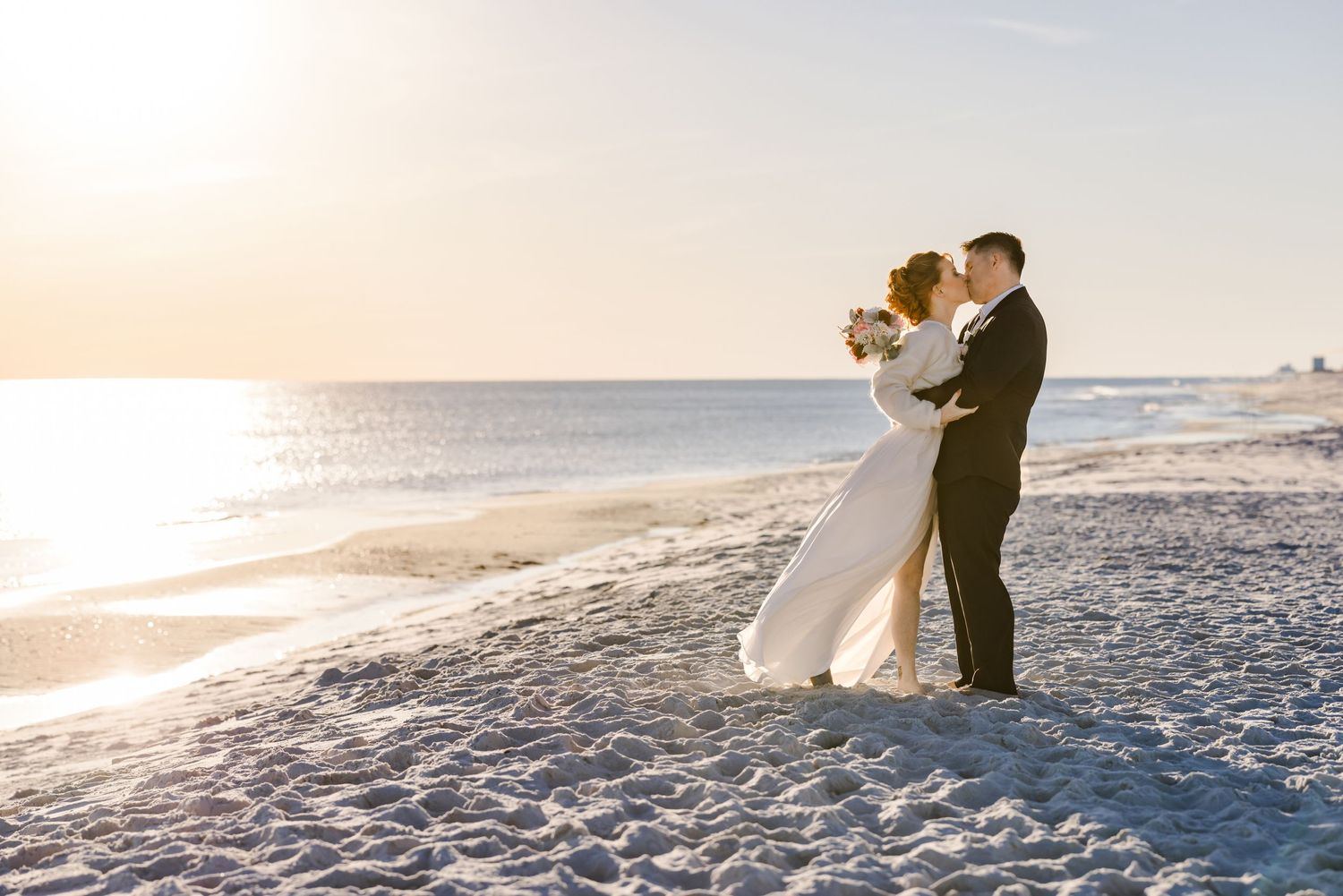 A couple in wedding attire embraces on a sandy beach at sunset during their romantic seaside celebration.